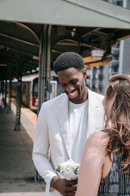A man in a white suit is smiling warmly while holding a bouquet of small white flowers wrapped in white paper, standing on an outdoor train station platform with a curved metal canopy overhead. His da