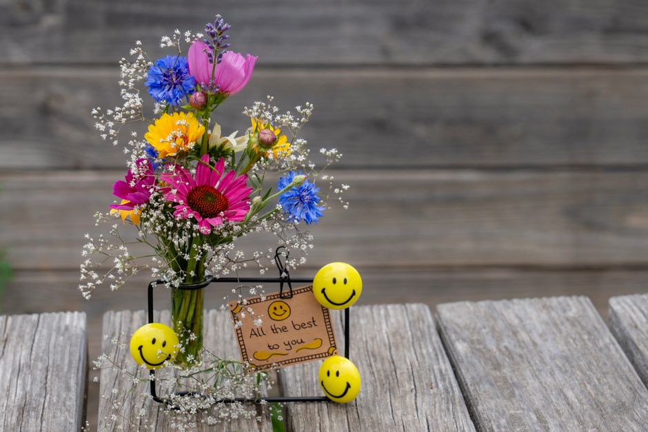 A vibrant bouquet of fresh flowers arranged in a clear glass vase, featuring pink gerbera daisies, purple carnations, blue cornflowers, yellow daisies, and delicate white baby's breath, positioned on